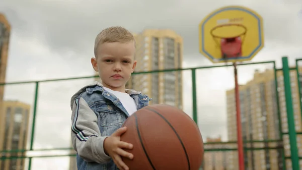 Junior schoolboy holds basketball in hands on sports ground against basketball hoop in dwelling district. Child dressed in stylish clothes close low angle shot