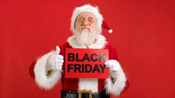 Mutlu Noel Baba, Kara Yazıtlı Afiş 'in maketini tutan Kara Cuma büyük indirimle seviniyor ve onaylayıcı bir parmak hareketi gösteriyor. Kara Cuma 'da büyük indirim var.