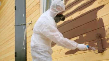 Man professional painter in protective suit respirator painting wooden house wall from outside using roller brush in brown color. Repair construction finishing works, final stage of home renovation.