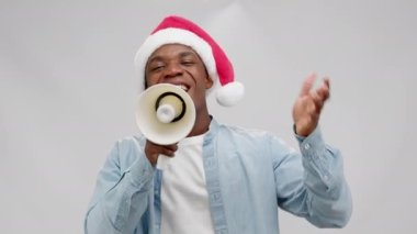 Young African-American man announces opening of annual Black Friday via megaphone. Cheerful guy in Santa hat speaks actively gesticulating with hand