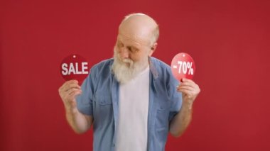 SALE -70 Off. Cheerful old man Dancing and Joyful From Black Friday Sale Holding Two Banners With Inscription SALE and -70 Off Showing Off Inscriptions to Camera on Red Studio Background