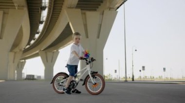 Little boy stands with bicycle under high overpass city bridge with small colorful windmill on handlebar. Junior schoolboy dressed in white T-shirt and denim shorts