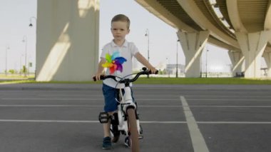 Junior bike rider in casual clothes stands turning head under overpass against city street lights and road signs. Serious boy has spinning windmill on handlebar