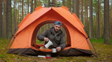 Camera zoom on traveling person man sitting in nature in tourist orange tent, pours himself a cup of hot fragrant coffee or tea and enjoys a drink among green trees Outdoor recreation, unity nature