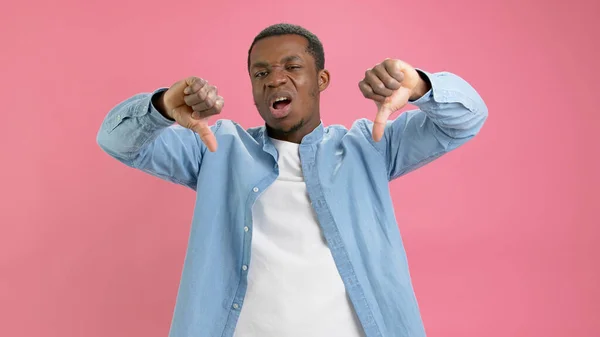 Disgruntled young black guy African American in denim shirt and white T-shirt shows thumbs down, dislike, negative feedback dislike, bad feedback, emotion of non-approval isolated on pink background