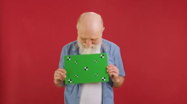 Happy old man holding Banner with Green Screen Tracks Points for Copy Space. Empty Green Screen Board. waves Head approvingly, approves of choice With Space for Text or Advertising on red Background