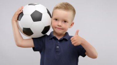 Little boy wearing blue t-shirt stands holding ball for playing football. Preschooler shows thumb-up with happy expression enjoying sports activities in white studio