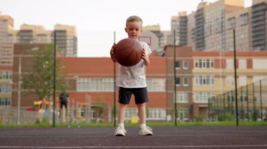 Happy preschooler wearing white t-shirt and black shorts bounces ball on empty basketball court. Boy enjoys playing basketball against orange and white buildings
