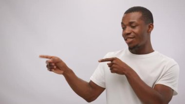 African American man does advertisement at white studio wall. Black performer with short haircut and beard in t-shirt advertises interesting stuff pointing fingers