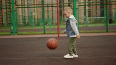 Child boy is practicing exersice hitting basketball ball training on court in city. Sports basketball skills concept. Kid is making his first steps as a player in basketball game. Outdoors urban life.