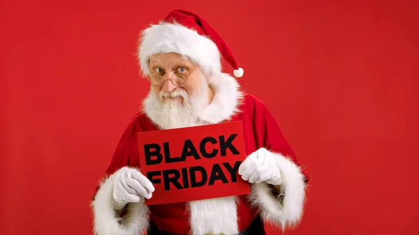 Sale Santa Claus on red Background holding lettering Black Friday in his hands and looking at camera. Merry Christmas sales from Santa Claus, Dancing with banner Black Friday sign and smiling