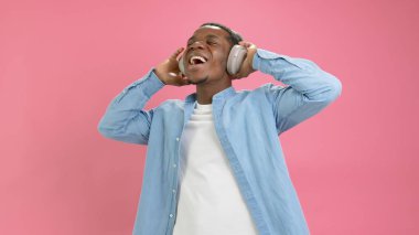 Portrait happy smiling African American man with grey headphones listening music and enjoying rhythm of song on pink background. Black person doing dance moves while using headset for entertainment
