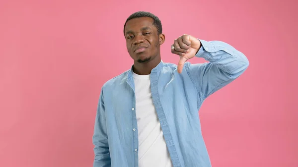 Not happy African American 20 years old in denim shirt, white T-shirt shows thumbs down, dislike, negative feedback, waves boy and says no, filmed in studio on pink background. High quality 4k footage