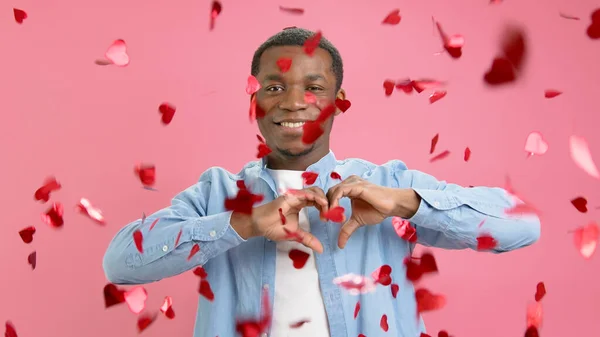 smiling African-American congratulates everyone on Valentines Day or Mothers Day, throws up confetti from red hearts, making heart gesture with fingers, showing love, in pink background
