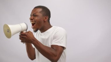 Young nervous black man with beard performs strange sketch at audition shouting to megaphone. African American man hands shake holding mouthpiece at white studio wall
