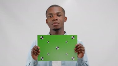 Afroamerican man with green screen banner in hands shakes his head negatively on white background. Empty green screen board with space for text or advertising. Concept of advertising and marketing.