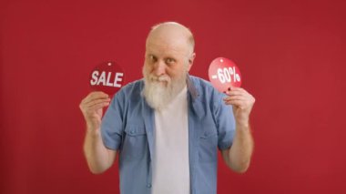 SALE -60 Off. Cheerful old man Dancing and Joyful From Black Friday Sale Holding Two Banners With Inscription SALE and -60 Off Showing Off Inscriptions to Camera on Red Studio Background