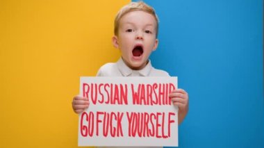 Portrait small blond boy raising banner with inscription shouts slogan Russian warship, go fuck yourself, standing on blue-yellow background of studio. No war, stop war, Russian aggression