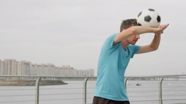 Man soccer player is rolling balancing football ball by his hands and back training on waterfront on city street. Footballer is doing exercises outdoors with ball in freestyle. Sports skills concept.