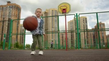 Child boy is hitting a basketball ball training on court practicing exersice. Sports basketball skills concept. Kid is making his first steps as a player in basketball game. Outdoors urban life.