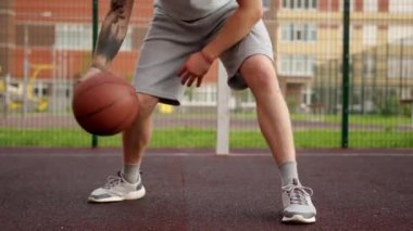 Sportsman with tattoos on hand demonstrates amazing tricks bouncing ball on basketball court. Adult wearing grey shorts and sneakers enjoys playing basketball