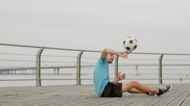 Man soccer player is performing amazing tricks with spinning football ball sitting on waterfront in the city. Footballer is training outdoors in freestyle. Improving honing sports skills concept.