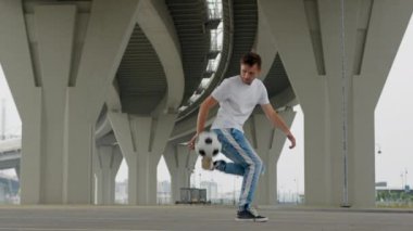 Man professional footballer is performing tricks with ball in freestyle training on city street under the bridge. Soccer player is practicing training outdoors juggling ball improving sport skills.