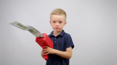 Little cute blondy boy shoots dollars from plastic red gun and smiling with happy face on white background. Funny time, childhood, marketing, advertizing concept. He is wearing blue polo shirt.
