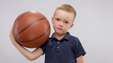 Portrait of smiling child boy showing thumb up be ine hands and holding basketball ball in other hand. Sport and active lifestyle for kids concept. Kid is wearing blue polo shirt.