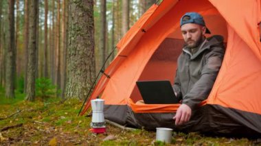 Bearded 25 year old man is working at laptop, drinking fragrant black coffee sitting in nature in an orange tent among wildlife and coniferous trees. Work outside office, outdoor recreation in spring