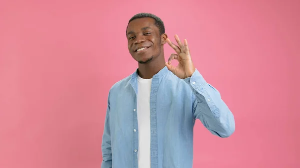 Excited funny 18 year old young bearded African American, dressed in denim shirt, looks at camera, speaks and gestures showing gesture OK posing in isolation on pink background in studio