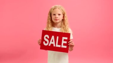 Cheerful blonde caucasian teenage girl in white T-shirt dances and smiles holding red banner with inscription sale on studio background. Girl advertises big sale on Black Friday and big discount