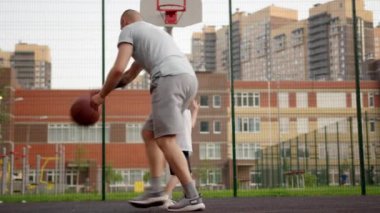 Father plays basketball circling around happy toddler son with ball on court. Excited preschooler runs laughing and smiling on basketball court against school