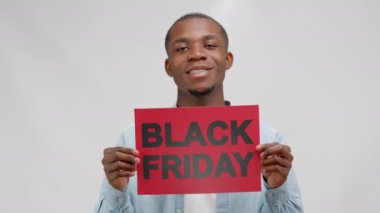 Afroamerican smiling man in denim shirt rising red banner black friday in hands on white background. Concept of sales, advertising and marketing. African american man on sale with black friday poster.