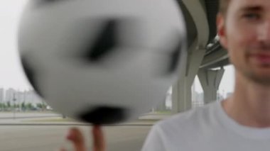 Portrait of soccer player in white t-shirt is spinning a ball on his finger. Footballer is training outdoors with ball in freestyle. Sports football skills concept. Young man performing a ball trick.