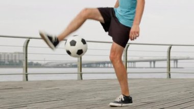 Professional man footballer is juggling a ball with his feet on waterfront, legs close-up. Soccer player is training outdoors with ball improving skills in slow motion. Sports football concept.