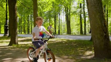Focused child boy is riding a bike with colourful pinwheel in city park in slow motion among trees at summer day. Outdoors activities for kids and happy childhood concept. Modern urban life of kid.