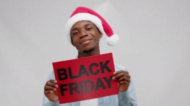 Happy chilling smiling dancing african american man in christmas hat holding banner with inscription black friday on white background and looking at camera. Concept of sales, advertising, marketing.