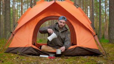 Bearded traveling man sitting in nature in tourist orange tent pours himself cup of hot aromatic coffee made on gas burner from white teapot and enjoys drink among green trees, Outdoor recreation