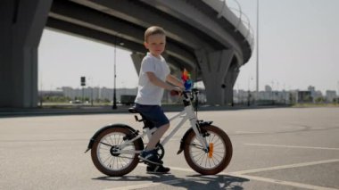 Outdoors activities for kids happy childhood concept. Child boy with bike is standing near the bridge on asphalt in the city at summer warm day. Camera is moving around the kid who looking at camera.