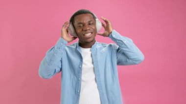 Portrait dancing black man on headphones. Slow motion young happy teenager 20 years old African American in denim shirt dancing listening to music with headphones on head on pink studio background