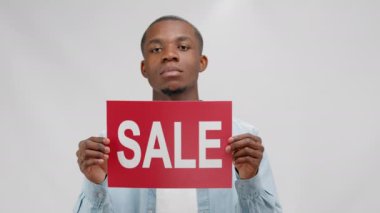 Serious young african american man holding red banner with inscription sale on white background. Sale message with place for text, copyspace. Concept of sales, advertising, black friday and marketing.