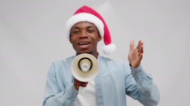 Laughing african american man in red christmas hat and blue denim shirt is speaking shouting loudspeaker on sale, black friday on white background. Concept of sales, advertising and marketing.