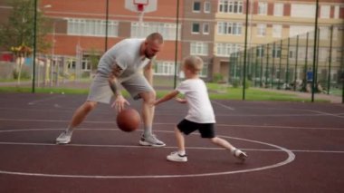 Father and son are playing together basketball on court in the city at summer day. Sports basketball skills and family time, parenthood concept. Outdoors urban life.