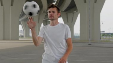 Young man professional soccer player is spinning a ball on his finger and looking at camera on city street. Footballer is training outdoors with ball in freestyle. Sports football skills concept.