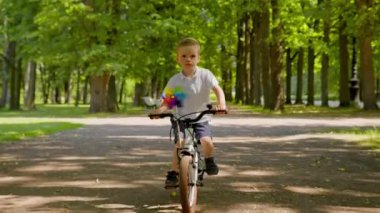 Focused child boy in white t-shirt and shorts is riding a bike in city park on asphalt road among trees at summer day. Outdoors activities for kids, happy childhood concept. Modern urban life of kid.