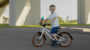 Serious child boy with bike is standing under the bridge on asphalt in city at summer warm day looking at camera. Outdoors activities for kids happy childhood concept. Camera is moving around kid.