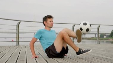 Young man soccer player in sportswear is practicing kicks with ball sitting on waterfront honing his technical skills. Professional footballer is training outdoors. Sports and football skills concept.