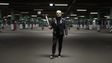 Halloween horror. Evil scary scarecrow with bag on his head in dark outfit walks through night parking lot and swings baseball bat. Masked maniac hunts victim at night on doomsday night
