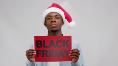Serious african american man in christmas hat holding red banner with inscription black friday on white background and looking at camera. Concept of sales, advertising and marketing.
